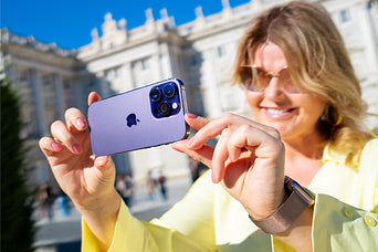 A woman smiling while using a refurbished purple iPhone 14 Pro to take a photo outdoors.