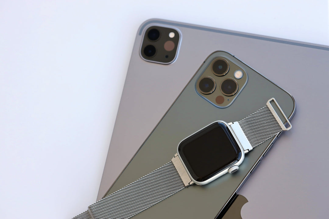A flat lay of high-quality refurbished Apple devices featuring an iPad Pro, an iPhone 12 Pro in Graphite, and an Apple Watch with a silver Milanese loop band.