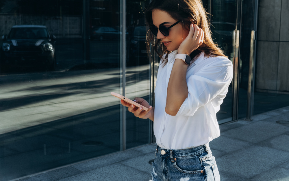 Brunette Woman In Sunglasses And White Shirt Walking Holding Pink Smartphone Wearing iWatch 